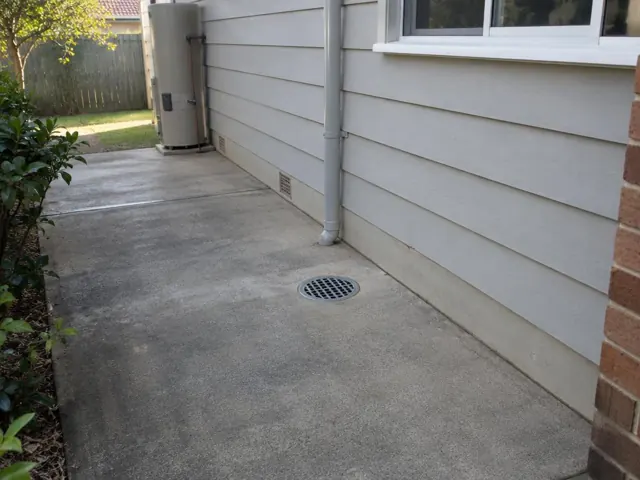 Outdoor concrete patio drain with grate beside a house wall and downpipe in Beacon Hill