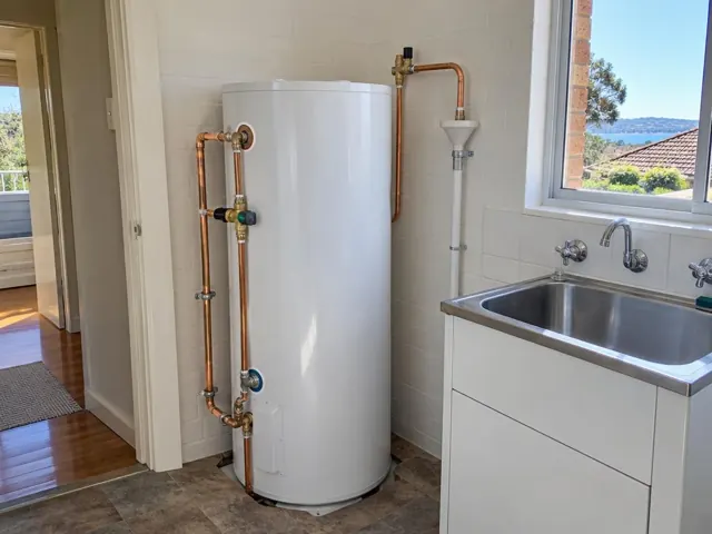 White hot water cylinder with copper pipework beside a sink in a tiled laundry room in Manly